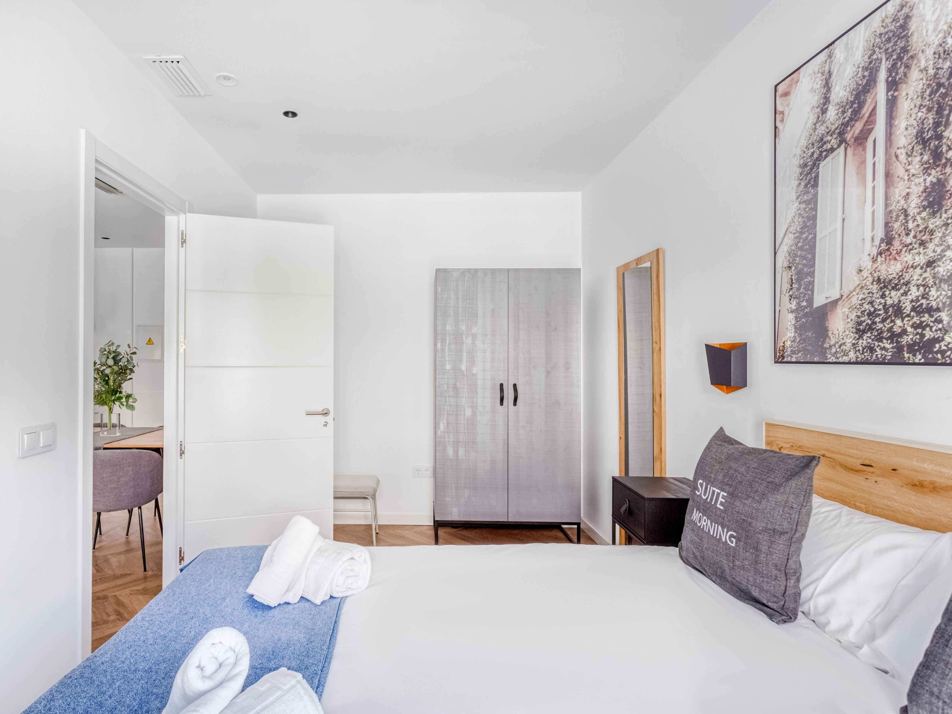 A modern hotel room with a bed, a bedside table, a mirror, a wardrobe, and a bathroom visible through an open door.