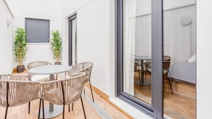 Un área de comedor exterior moderna con una mesa redonda y sillas de mimbre, flanqueada por plantas en macetas, frente a una pared blanca con un amplio ventanal corredera que da a una habitación contigua.
