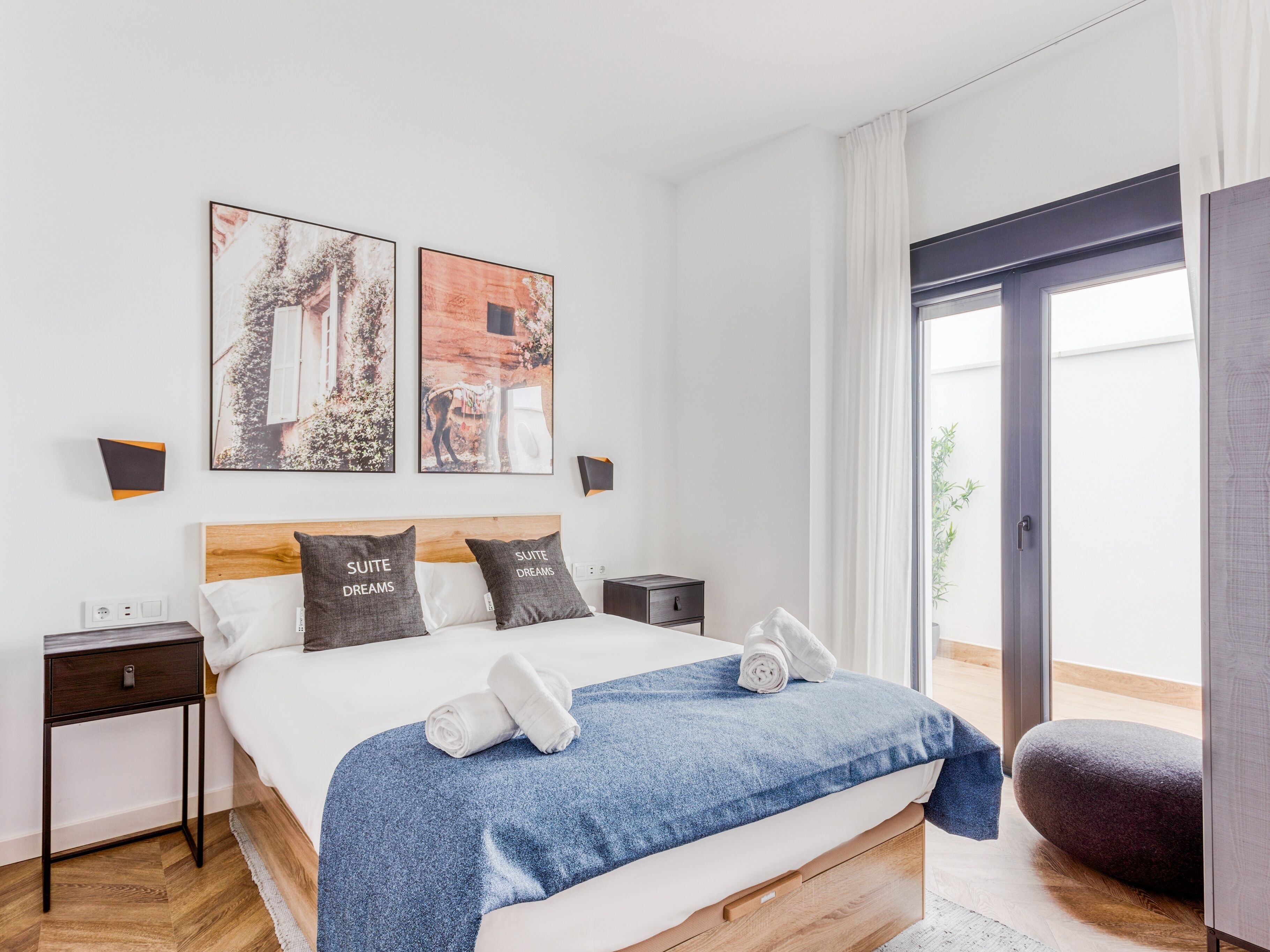 Un dormitorio moderno con cabecera de madera, una cama grande con manta azul, dos mesitas de noche y un ventanal con vistas a zonas verdes.