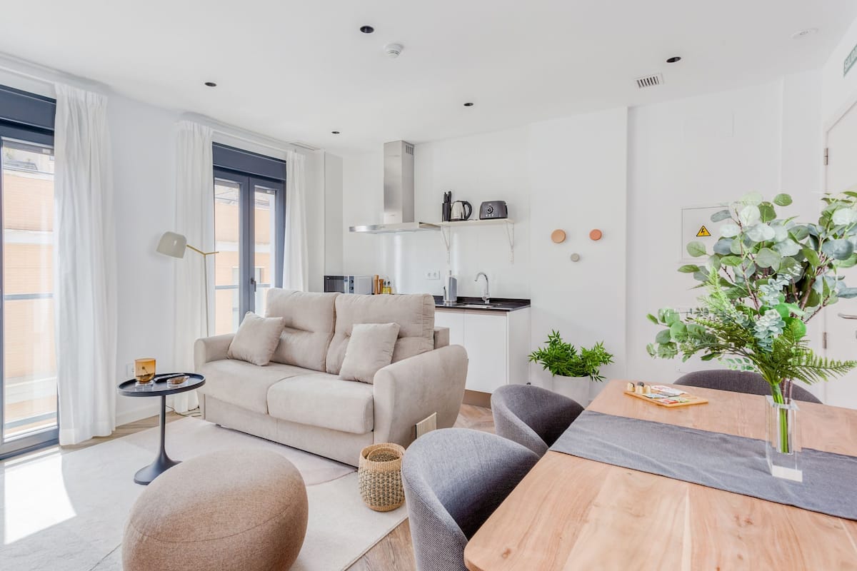 Ein modernes Wohnzimmer mit einer Couch, einem Esstisch mit Stühlen, einem kleinen Beistelltisch und einem großen Fenster mit Vorhängen.