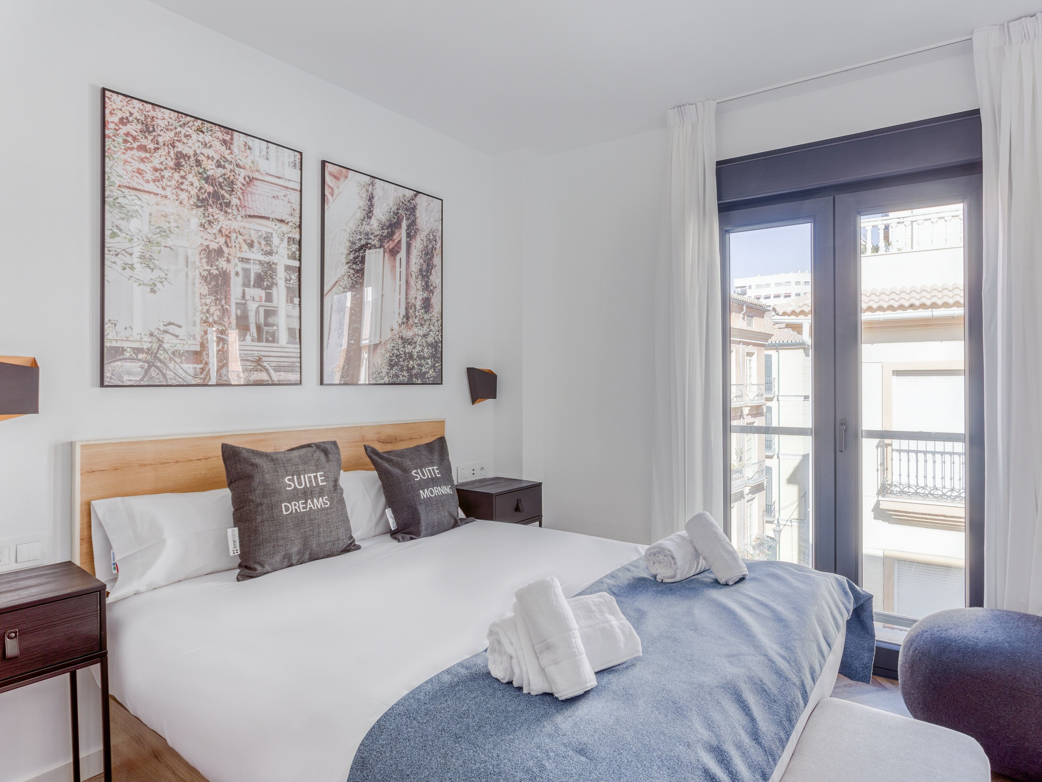 Een hotelkamer met een groot bed, twee nachtkastjes, een raam met uitzicht op gebouwen en twee kunstwerken aan de muur.