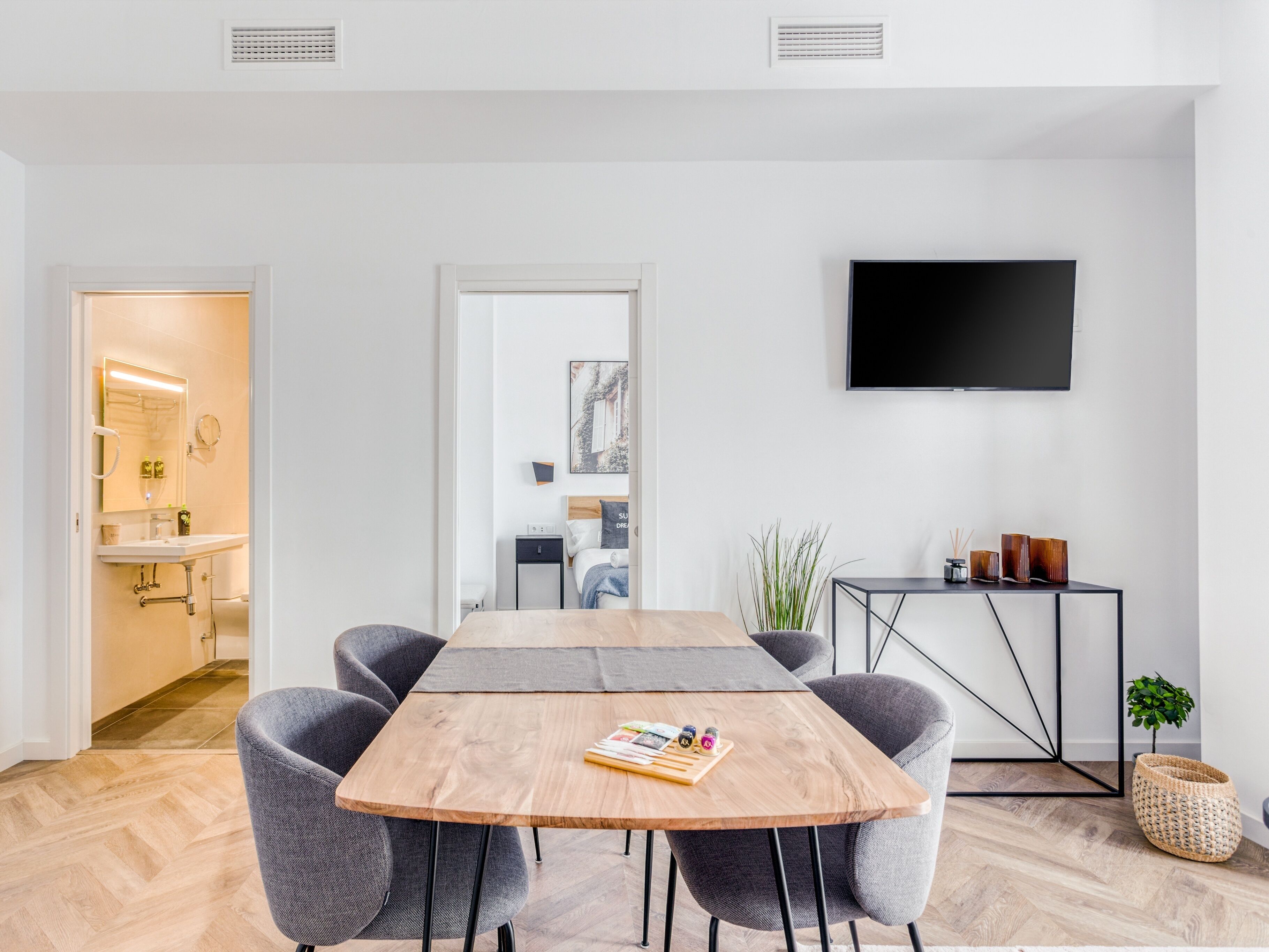 Een moderne hotelkamer met een houten eettafel, grijze bekleden stoelen, een flatscreen televisie aan de muur en een bijzettafeltje met decoratieve objecten.