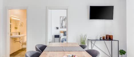 Een moderne hotelkamer met een houten eettafel, grijze bekleden stoelen, een flatscreen televisie aan de muur en een bijzettafeltje met decoratieve objecten.