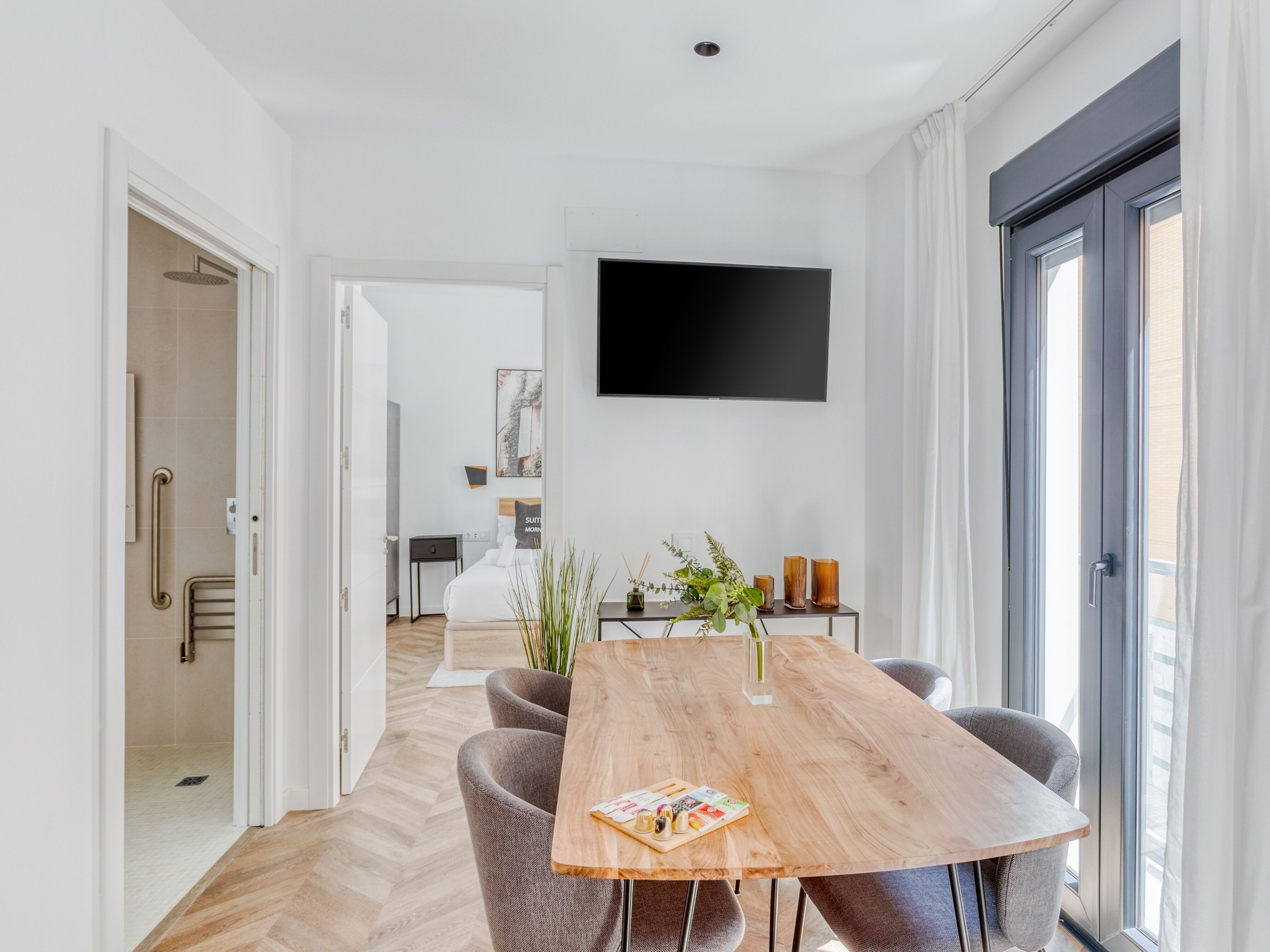 A modern dining area with a wooden table, grey upholstered chairs, a flat-screen TV mounted on the wall, and a view of the outdoors through a large window.