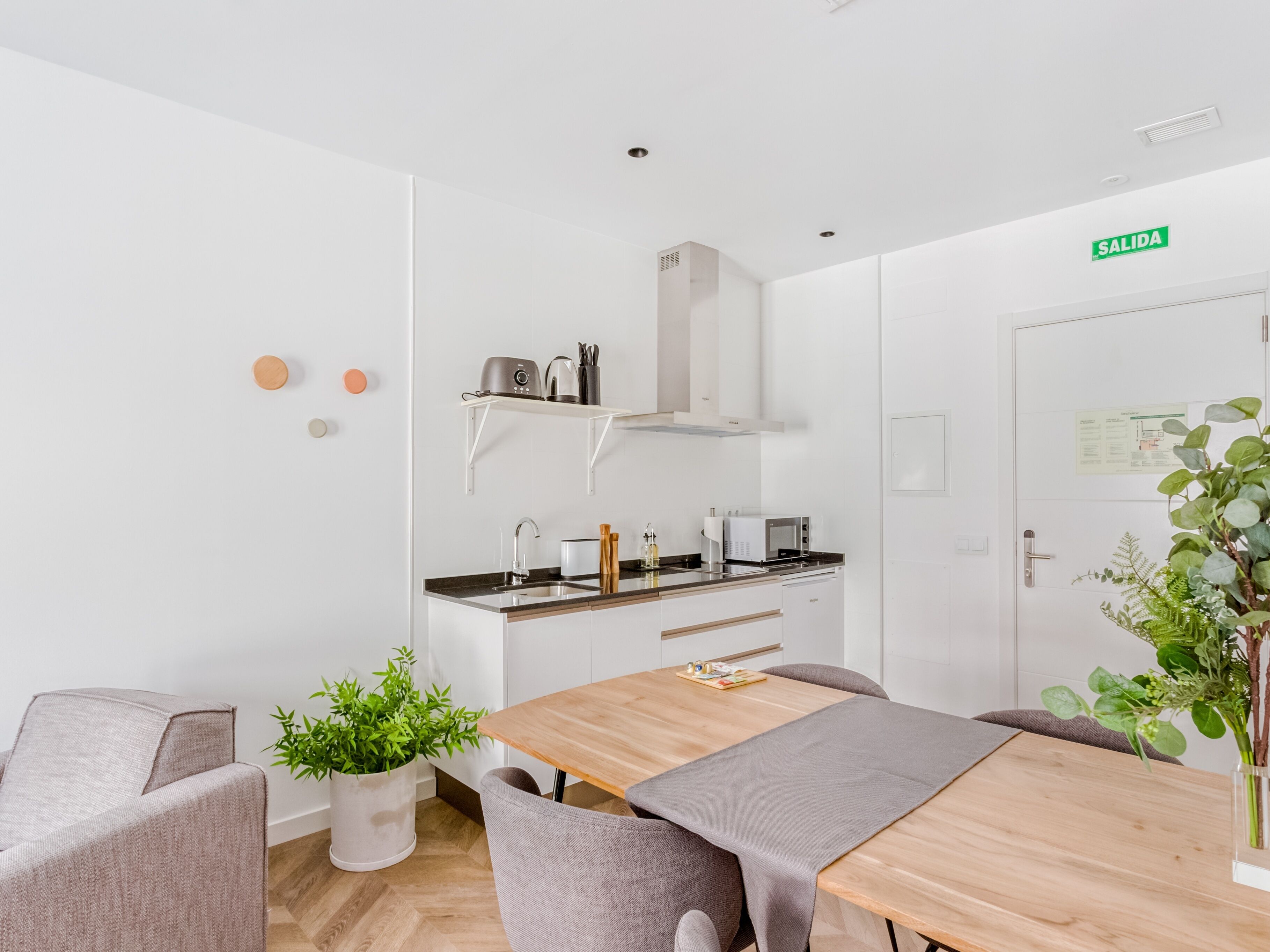 A modern kitchen with white cabinets, a wooden dining table, and a built-in microwave. There is a white door with a green 'SALIDA' sign, a potted plant, and a small shelf with kitchen utensils.