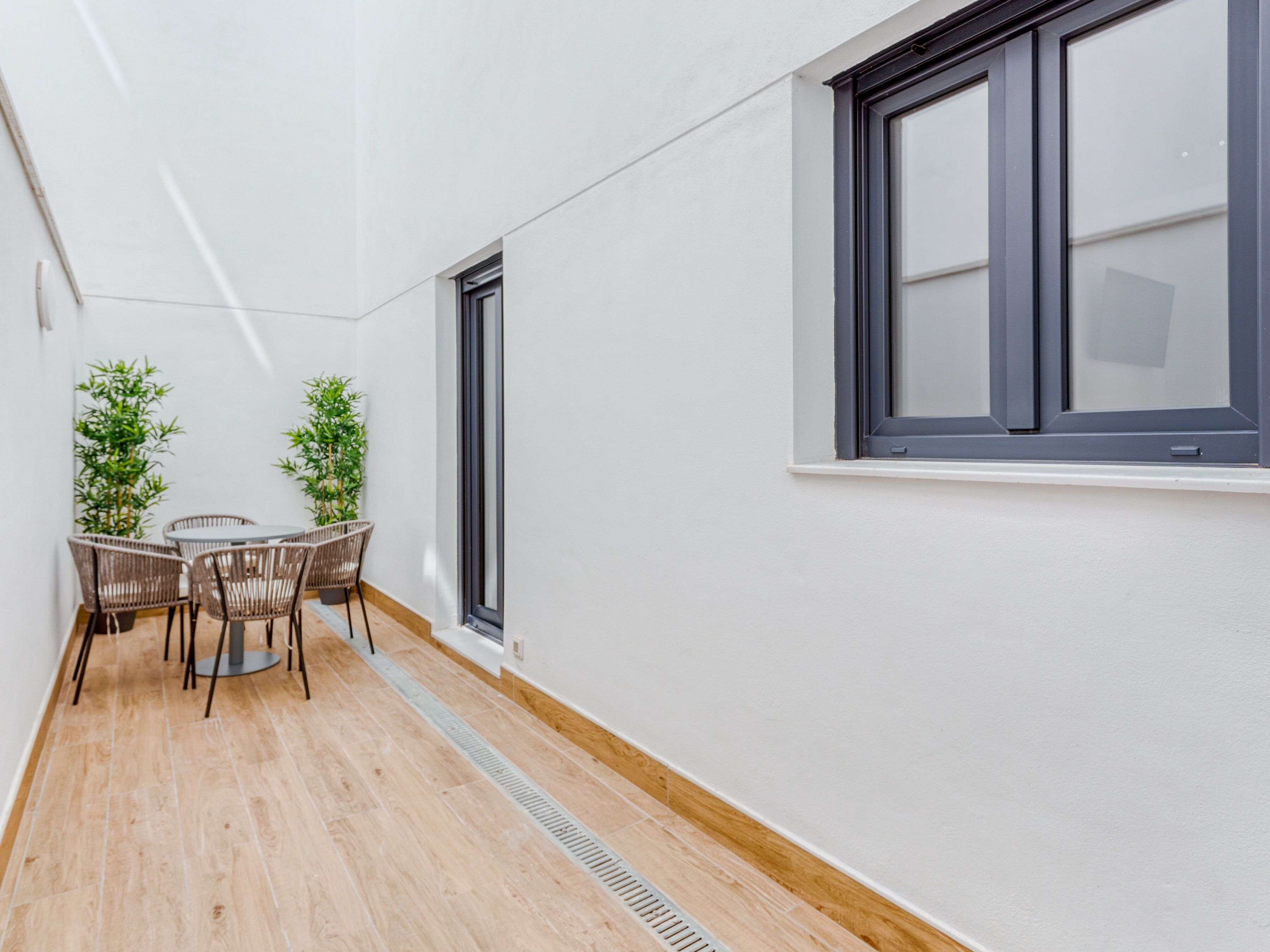Un estrecho pasillo con un rincón de comedor que incluye sillas de mimbre y una mesa, una ventana con marcos oscuros y una pared blanca.