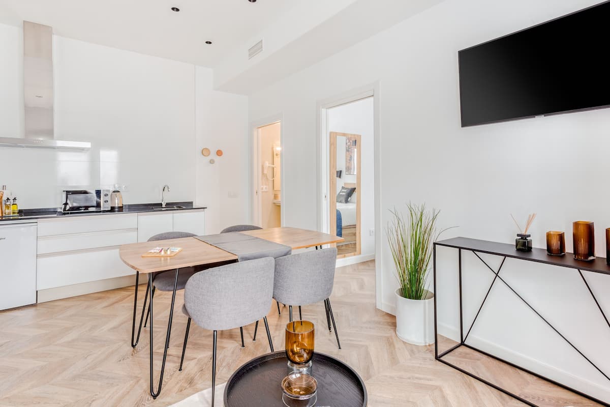 Eine moderne Küche mit weißen Schränken, ein Essbereich mit einem Holztisch und grauen Stühlen, ein an der Wand montierter schwarzer Fernseher und eine Topfpflanze seitlich.