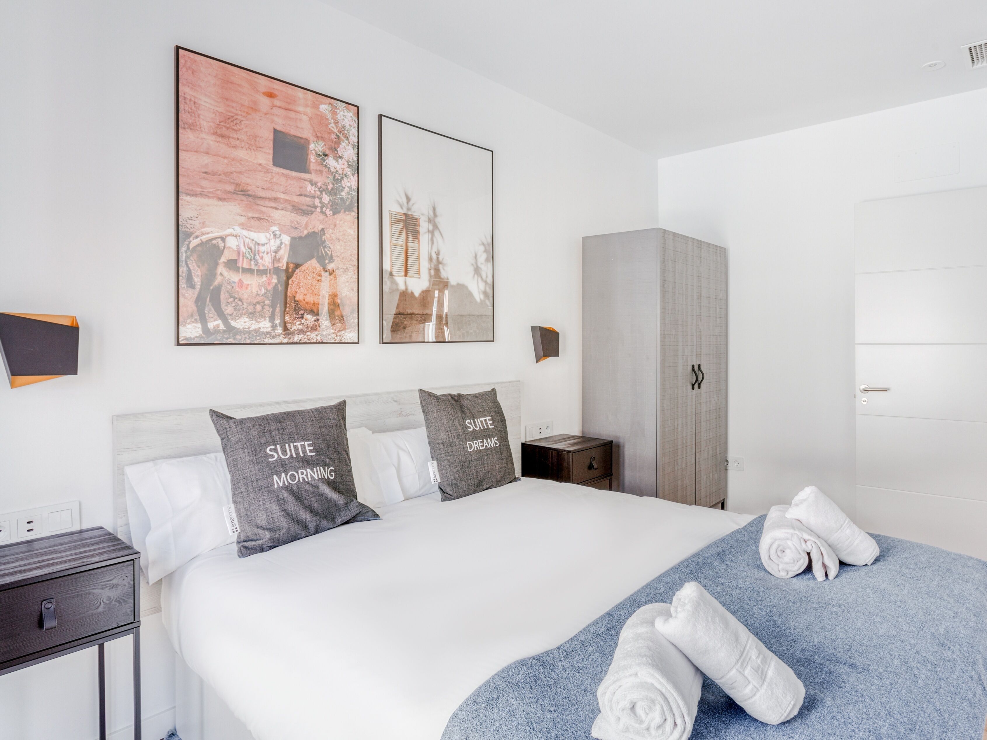 A modern hotel room with a large bed, two pillows with text, a wardrobe, and artwork on the wall.