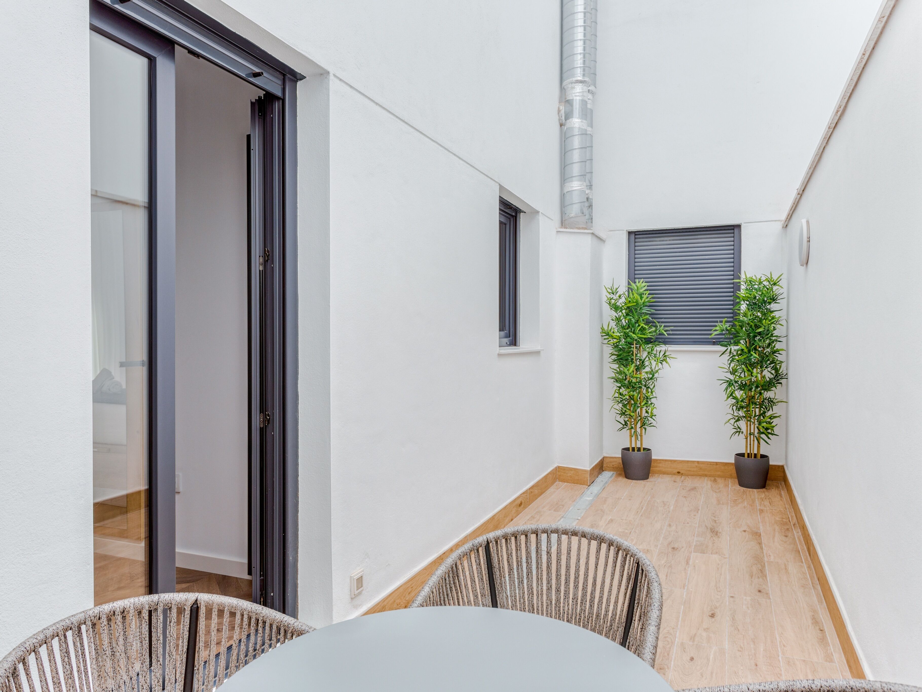 Un couloir étroit avec une porte coulissante en verre donnant sur une pièce, deux plantes en pot, une fenêtre avec des stores et une petite table avec des chaises.