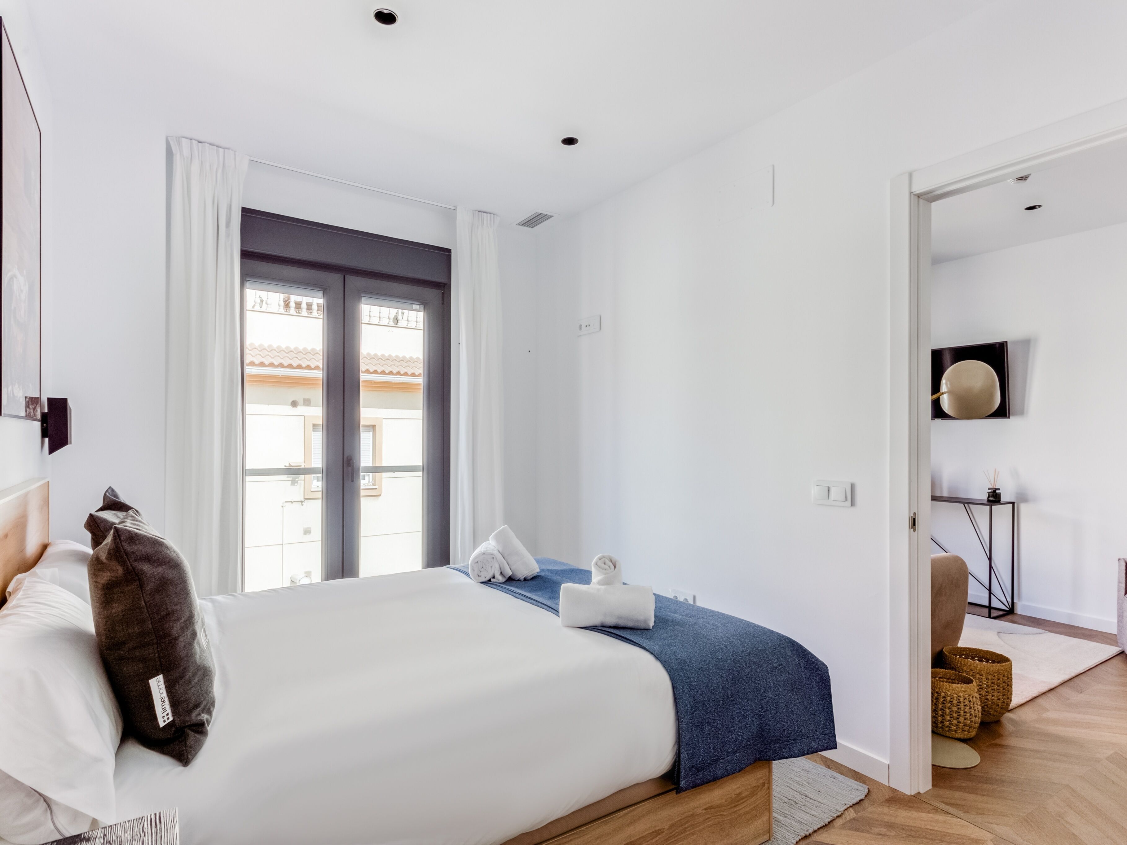 Une chambre moderne avec un grand lit, une fenêtre avec des rideaux, une table de chevet avec une lampe et une vue sur l’extérieur.