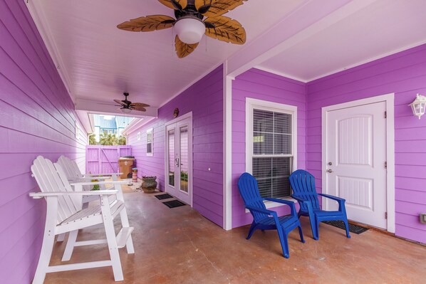 Eine Veranda mit weißen Adirondack-Stühlen und einem Tisch, ein violettes Haus und eine weiße Tür.