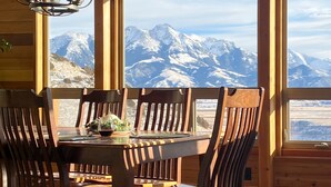 Una mesa de comedor de madera con seis sillas, una araña y vistas a montañas nevadas a través de amplios ventanales.