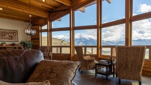 Una habitación acogedora con un sofá de cuero, sillones de mimbre y una mesa llena de libros. Amplias ventanas ofrecen vistas a las montañas.