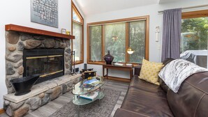Una sala de estar con chimenea de piedra, una mesa de centro de vidrio, un sofá de cuero y ventanas grandes con persianas.