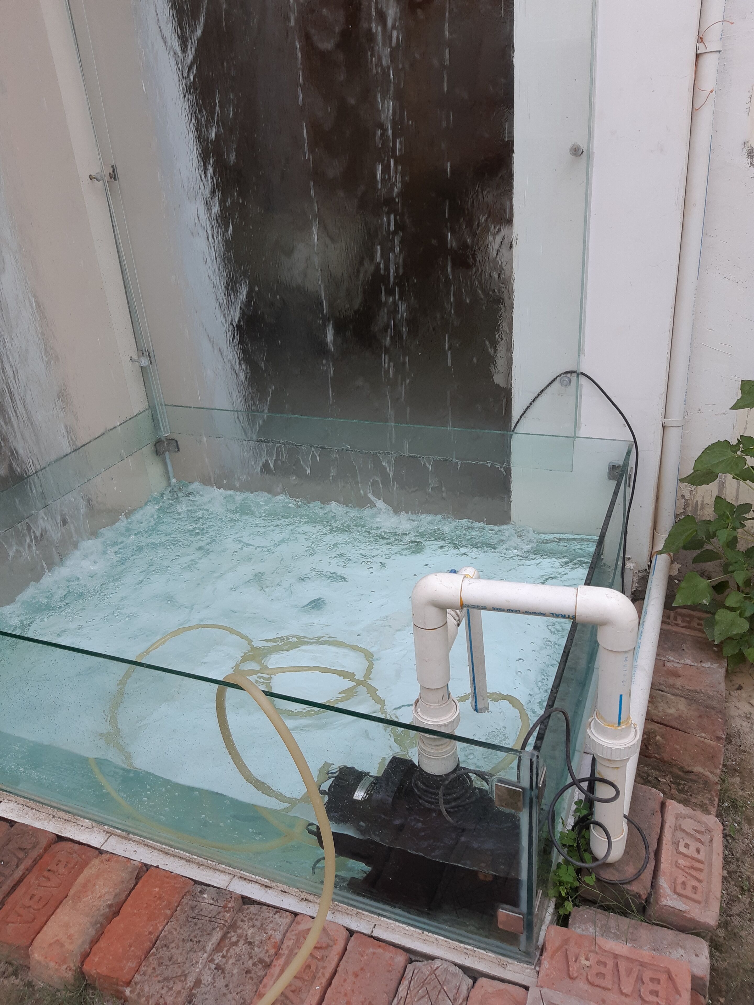 A glass-enclosed water feature with a pump and pipes, set against a brick wall.