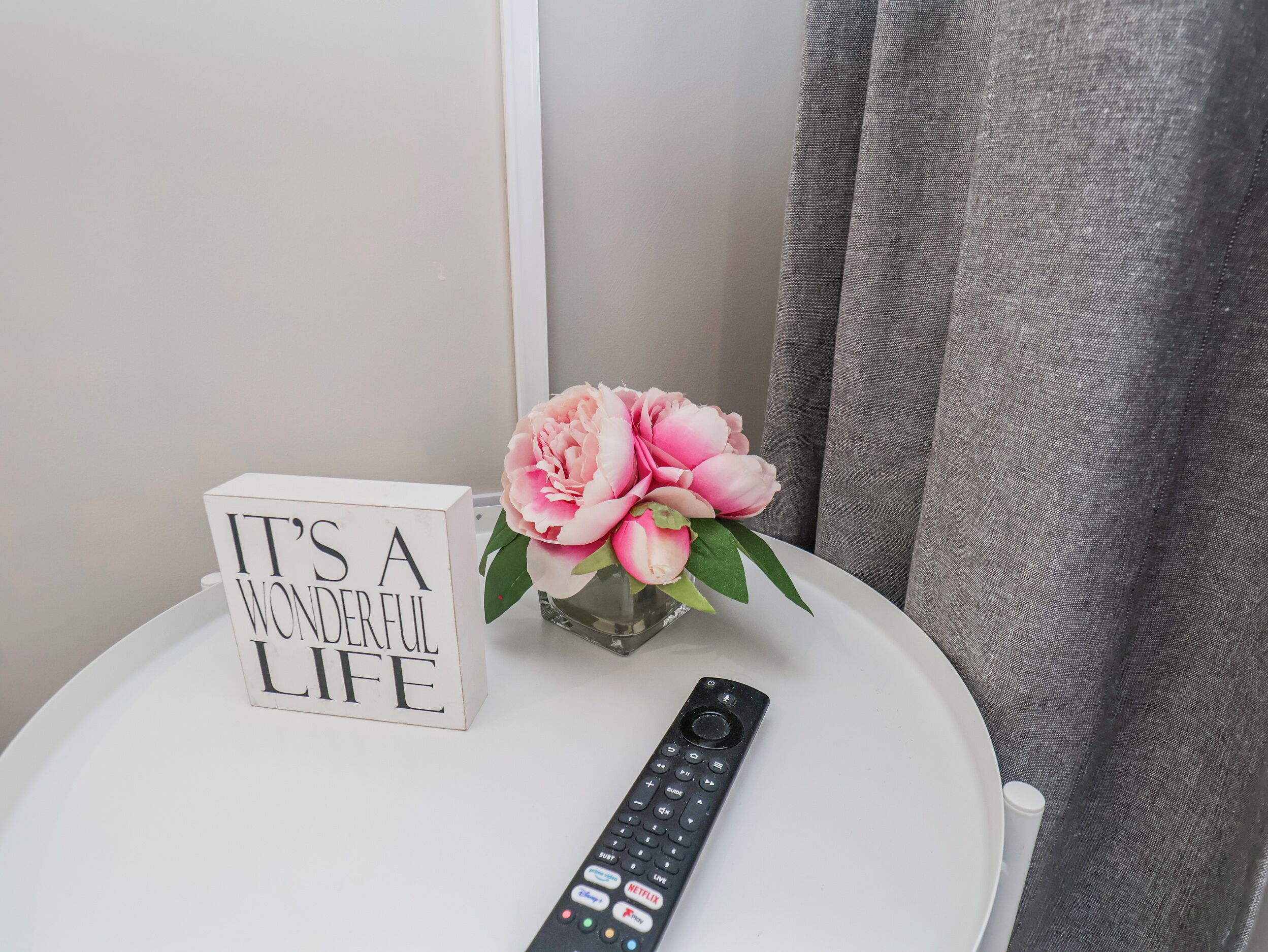 A white table with a sign reading 'IT'S A WONDERFUL LIFE,' a vase with pink flowers, and a remote control.