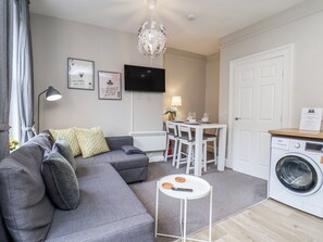A modern living room with a grey sofa, a dining table with chairs, a TV mounted on the wall, a washing machine, and a small round table with a remote control.