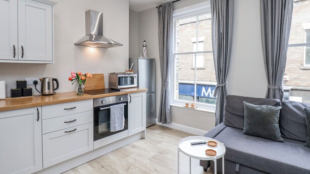 A compact kitchen with a microwave, kettle, and a small oven. There's a sofa with cushions and a small table with a remote control.