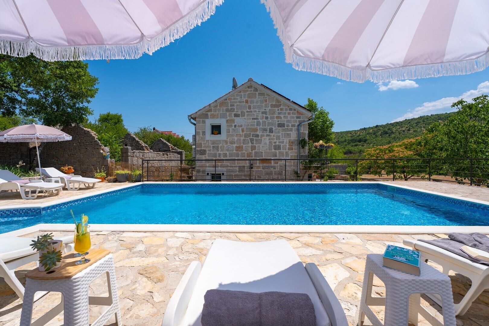A stone house with a swimming pool, surrounded by lounge chairs and umbrellas.