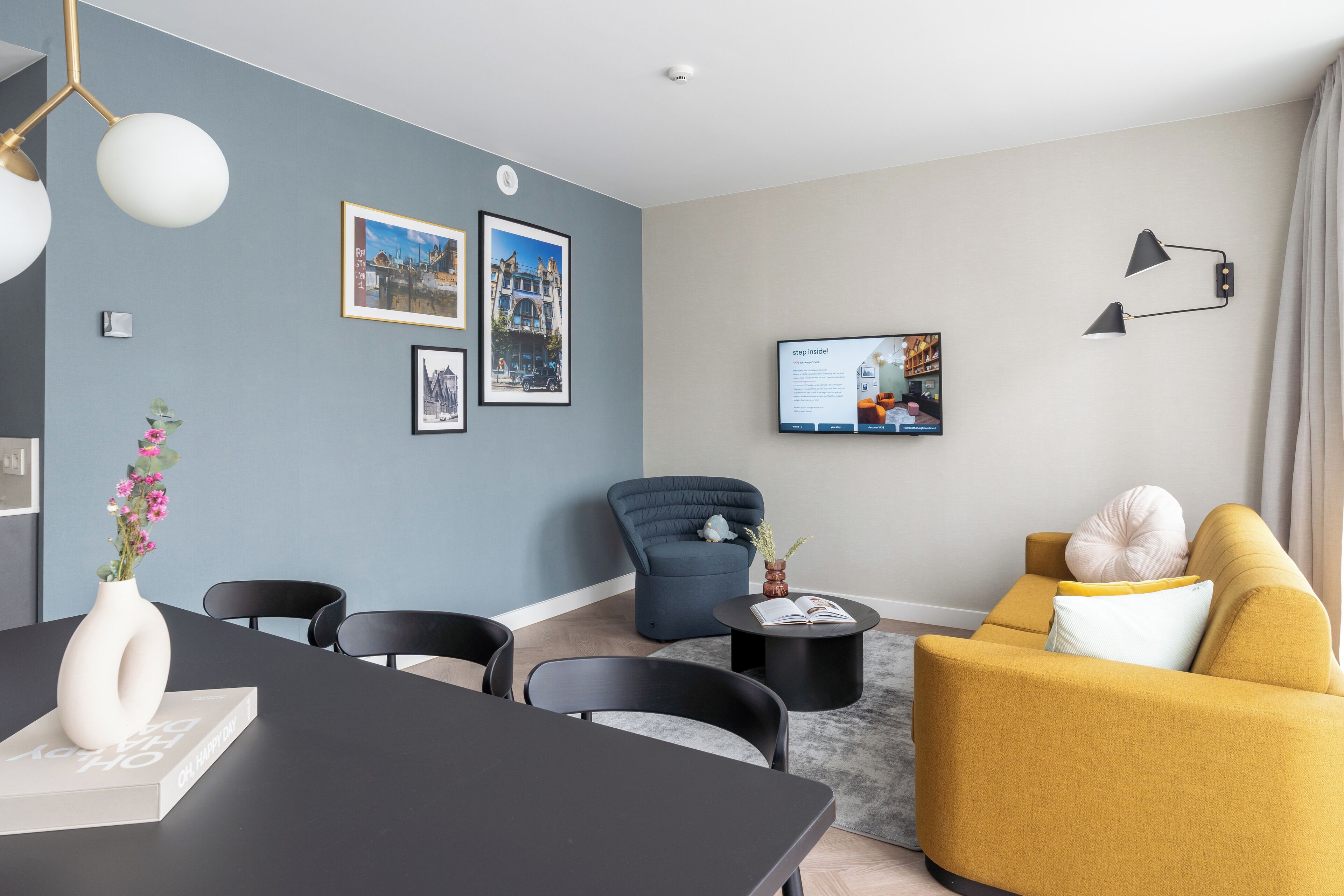 A modern living room with a dining area, featuring a sofa, chairs, a coffee table, and a flat-screen TV mounted on the wall.