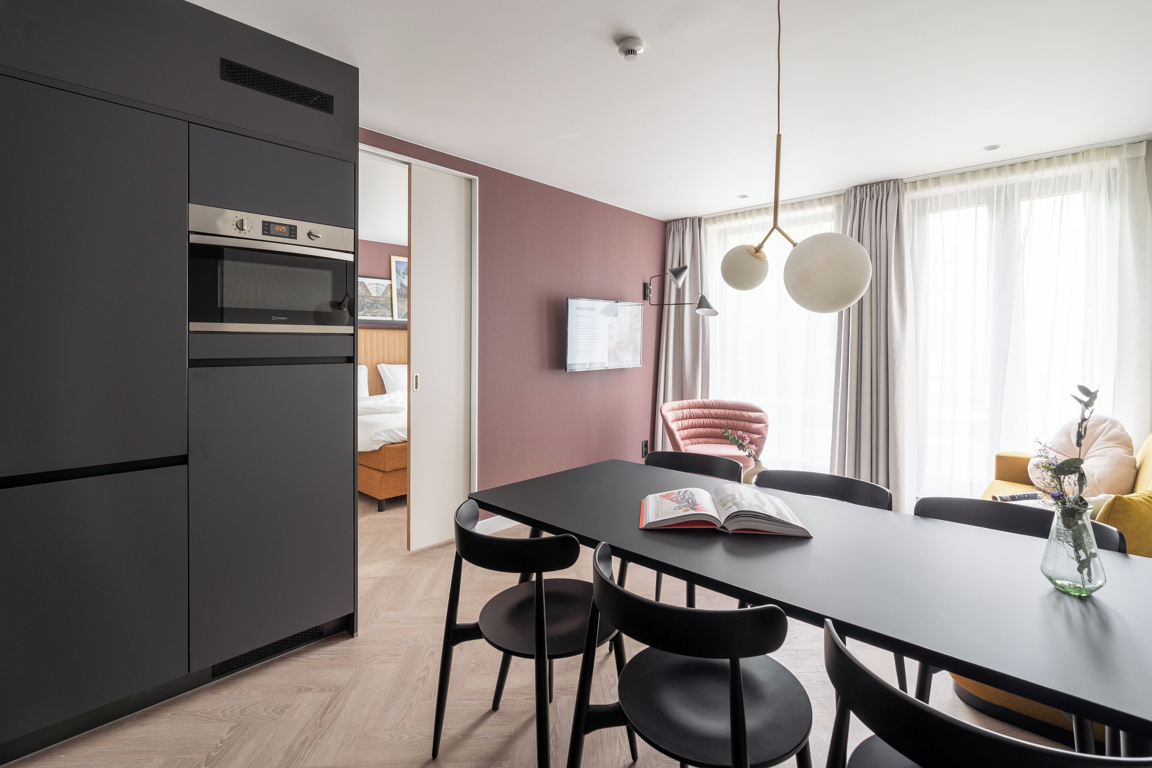 Eine moderne Küche mit integrierten Geräten, ein Essbereich mit Tisch und Stühlen sowie ein Blick auf ein Schlafzimmer durch eine offene Tür.