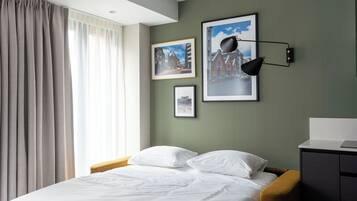 A neatly made bed with white linens, a wooden headboard, and a mustard-colored bench. There are framed pictures on the wall and a black desk lamp.