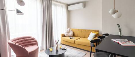 A modern living room with a pink chair, a yellow sofa, a round coffee table, and a dining table with chairs.