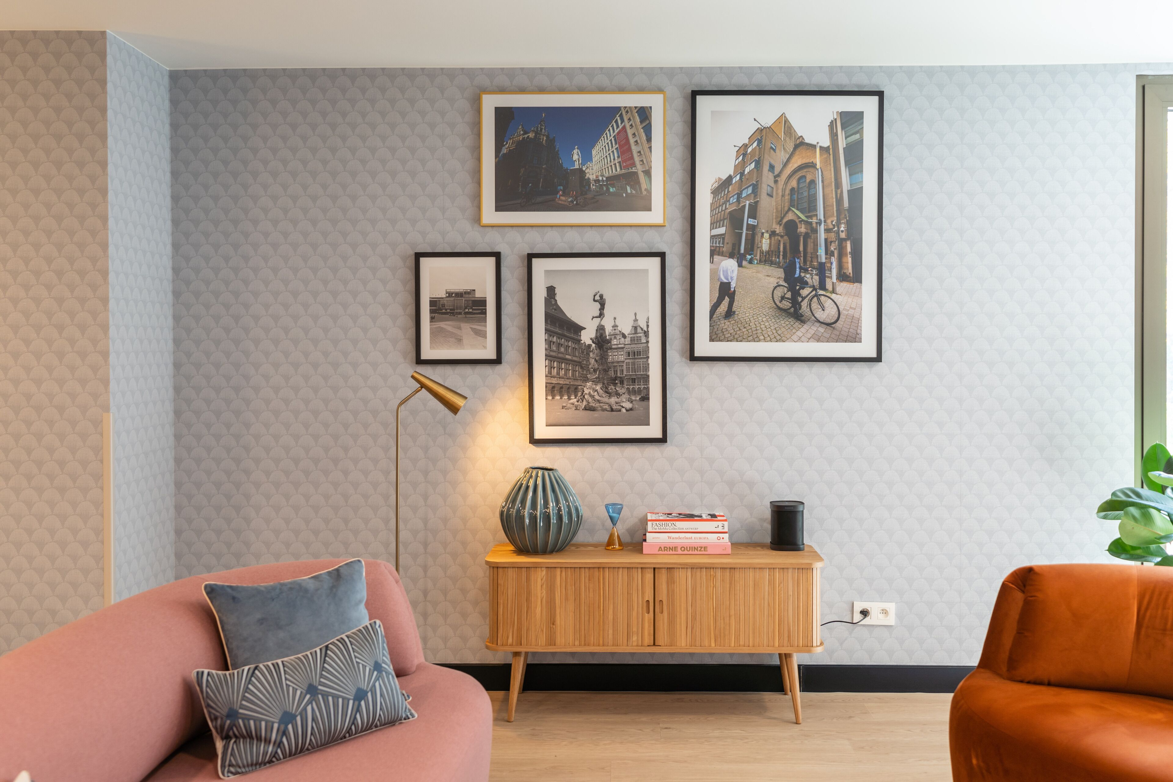 A living room with a sofa, a wooden side table with books, a floor lamp, and framed pictures on the wall.