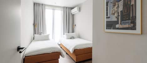 A hotel room with two beds, a window with curtains, an air conditioning unit, and a framed picture on the wall.