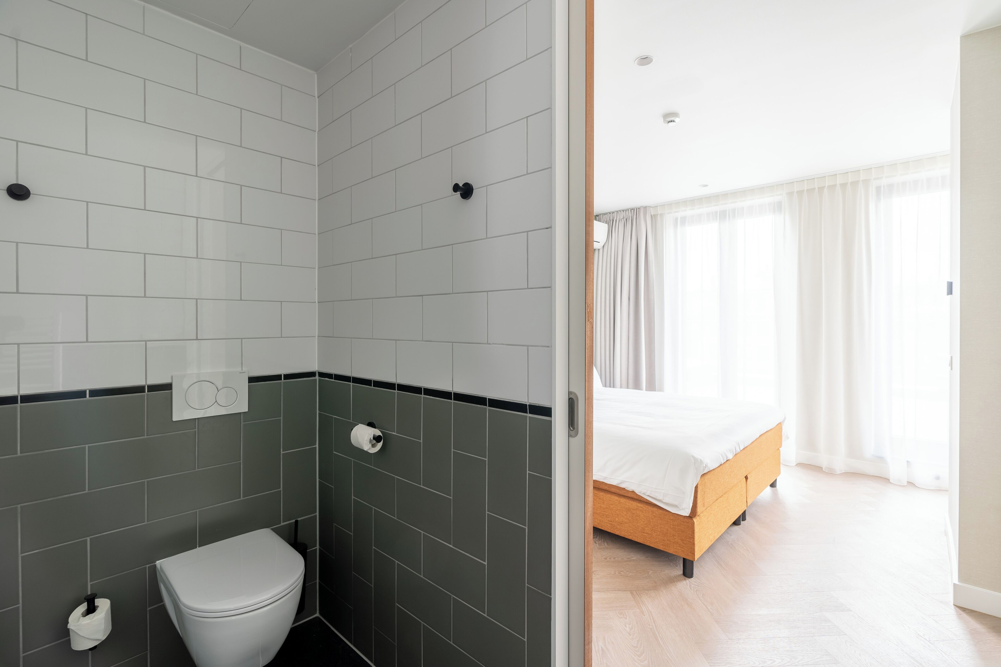 Ein modernes Badezimmer mit weißen und dunkelgrauen Fliesen, einer Toilette und einer Dusche. Angrenzend ein Schlafzimmer mit einem Bett und einem großen Fenster mit Vorhängen.