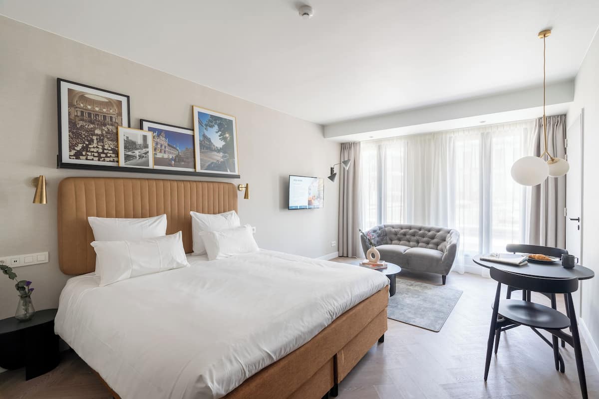 Habitació d'hotel amb un llit gran, un sofà armchair gris, una taula rodona petita i una finestra amb cortines.