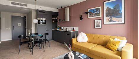 A modern living room with a yellow sofa, a dining table, and a kitchen area in the background.