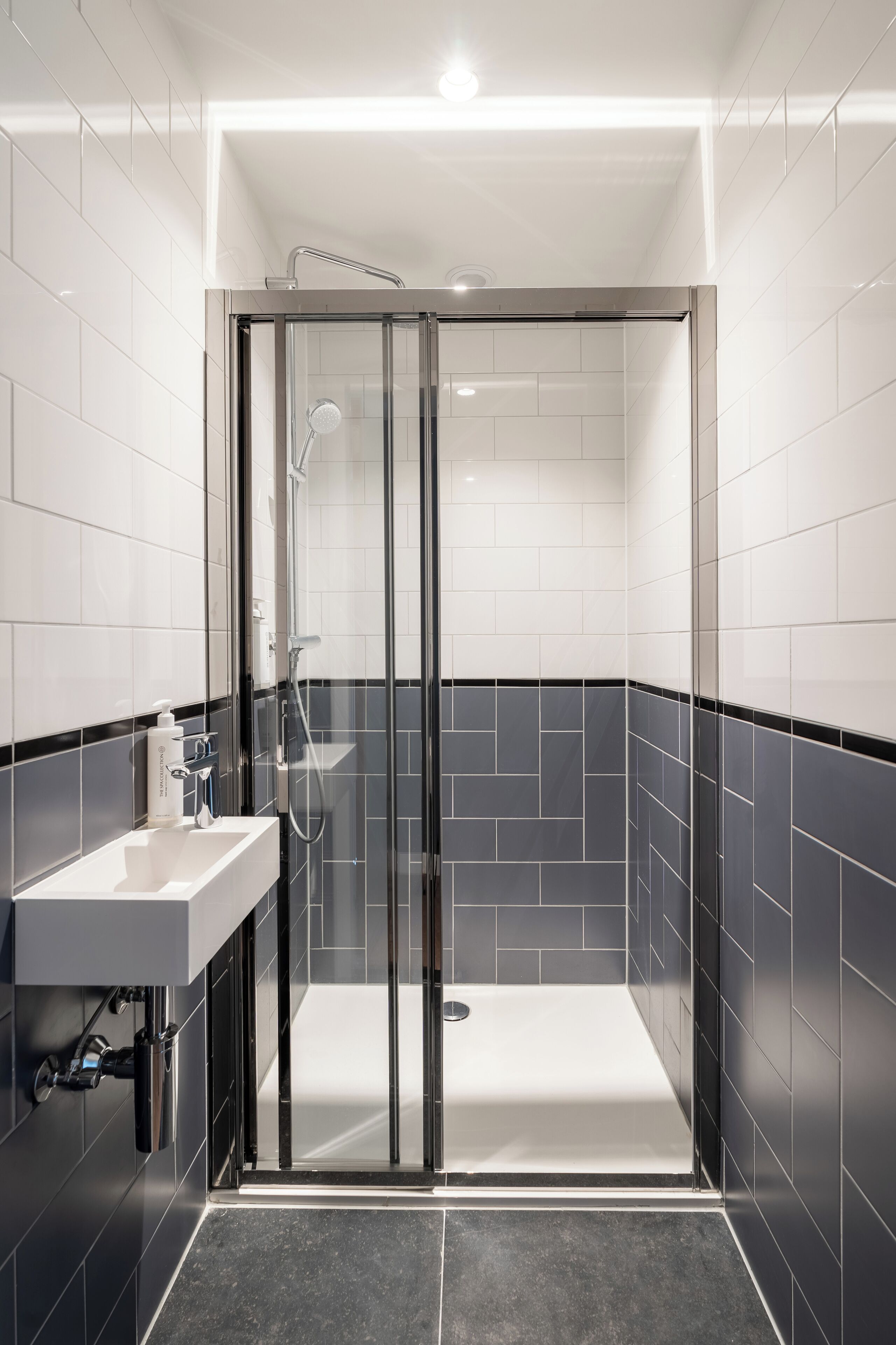Un bany modern amb dutxa de vidre, un lavabo blanc i parets revestides de rajoles negres.