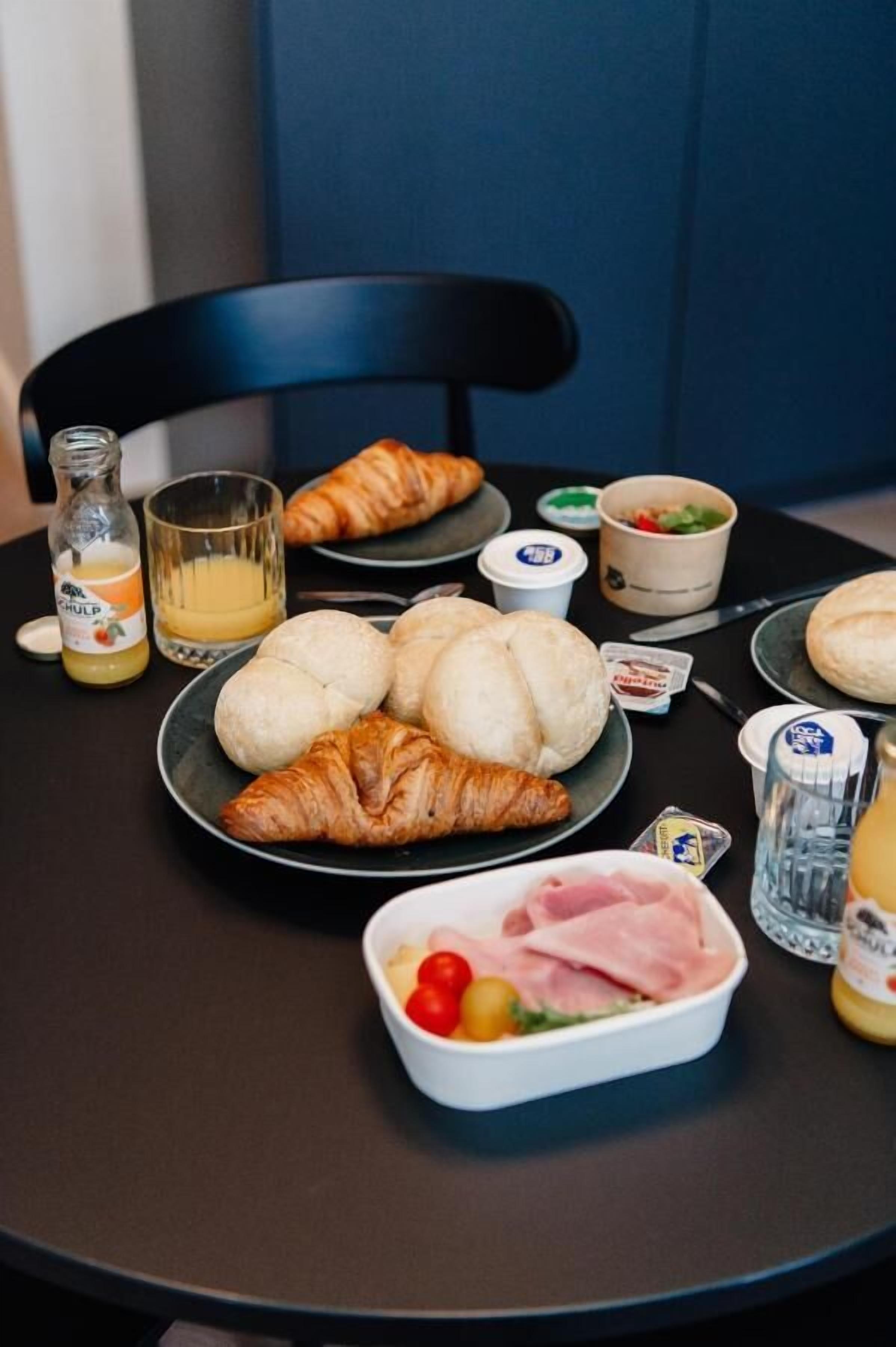 A breakfast table with croissants, bread rolls, ham, tomatoes, and a bottle of orange juice.