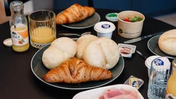 A breakfast table with croissants, bread rolls, ham, tomatoes, and a bottle of orange juice.