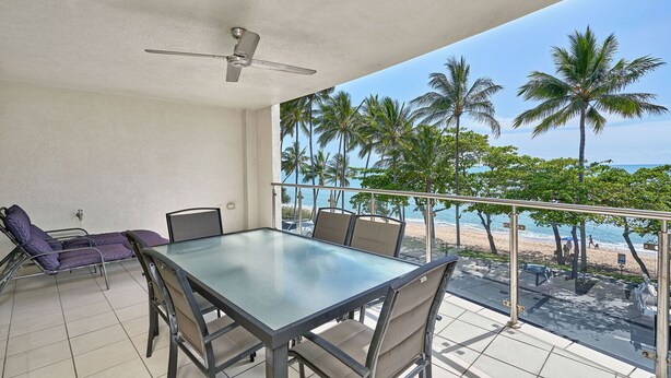 Un balcon avec une table et des chaises en verre, donnant sur une plage bordée de palmiers et sur l’océan.
