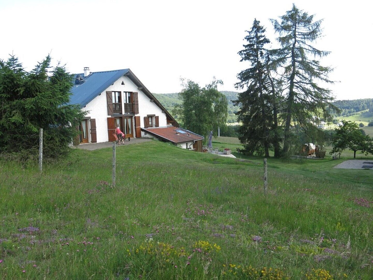 Une maison blanche avec un toit bleu, une terrasse en bois et un grand arbre en arrière-plan.