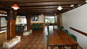 Un coin repas rustique avec une longue table en bois et des chaises vertes, des poutres en bois au plafond et une vue sur l’extérieur grâce à de grandes fenêtres.