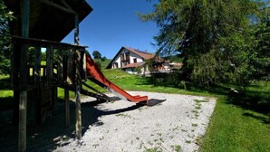 Une aire de jeux avec un toboggan et une structure en bois, entourée d’une pelouse et d’arbres.