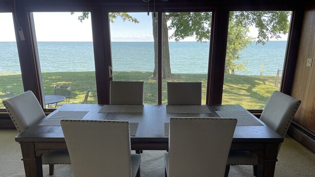 A dining area with a table and chairs, large windows with a view of the sea, and a grassy lawn outside. - Lake views and Sunsets for Days (Lorain)