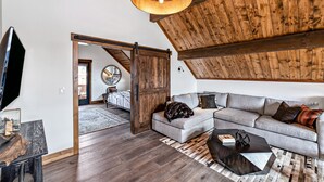 A spacious loft with a high ceiling, exposed wooden beams, a large gray sectional sofa, and a modern chandelier. - Slice of Heaven just steps from Lake Estes! Indoor/Outdoor Fireplace and Jacuzzi (Estes Park)