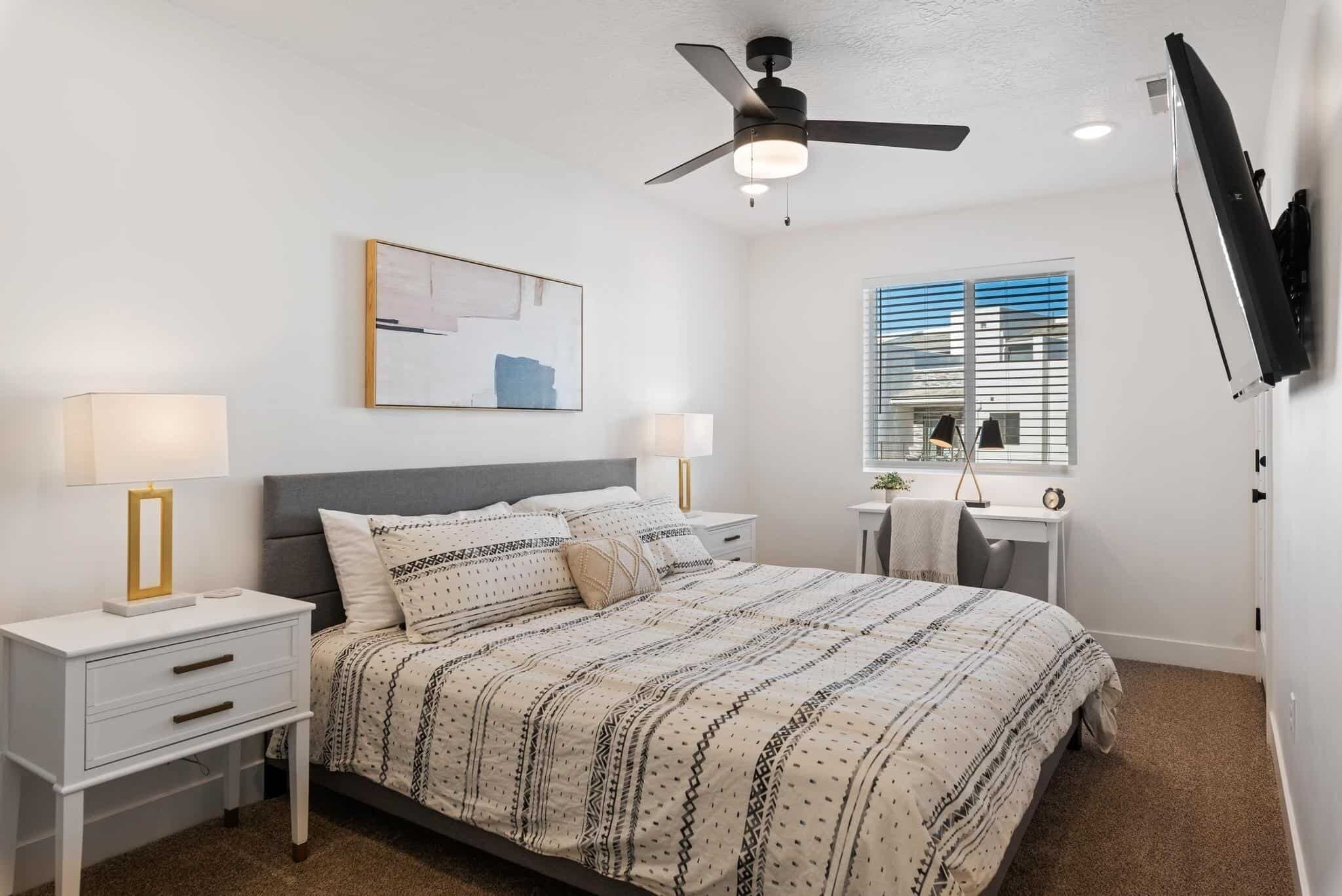 Une chambre à coucher comprenant un lit, des tables de chevet, un bureau, un ventilateur de plafond, une télévision et une fenêtre avec des stores.