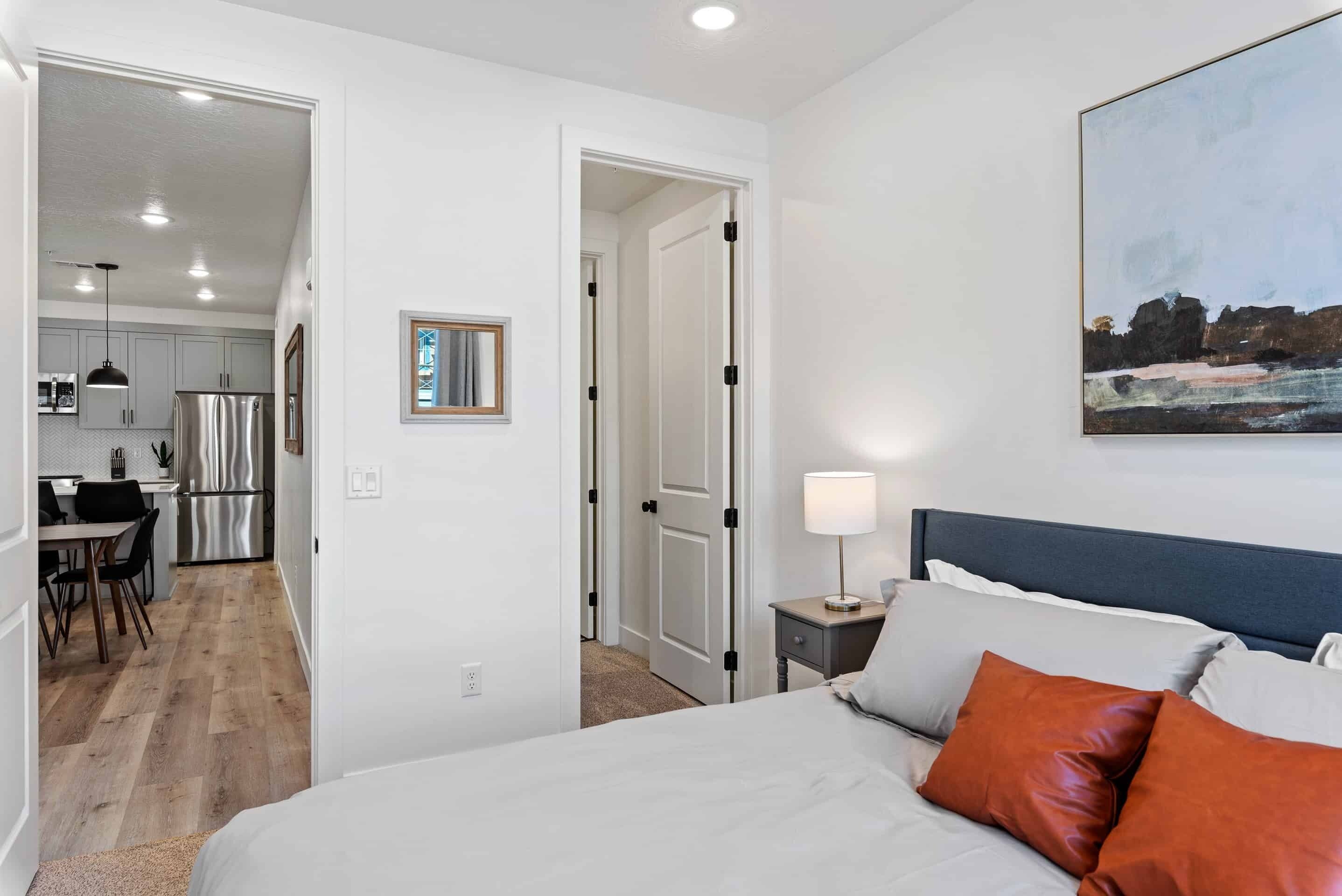 A bedroom with a bed, pillows, a nightstand, a painting, and a view of a kitchen and dining area.