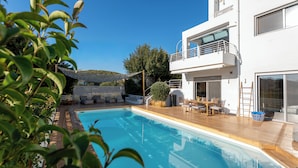 Un área moderna junto a la piscina con una terraza de madera, una piscina y un edificio blanco con balcón.
