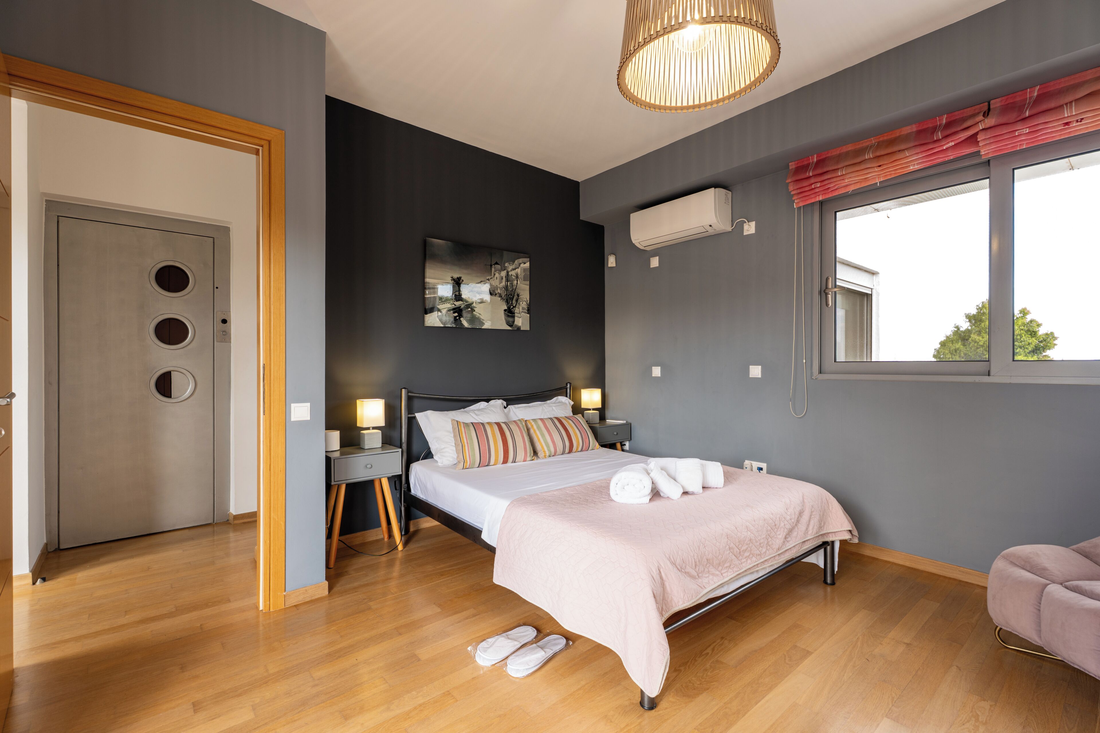 A modern bedroom with a bed, bedside tables, a wall-mounted air conditioner, a window with curtains, and a door with circular cutouts.