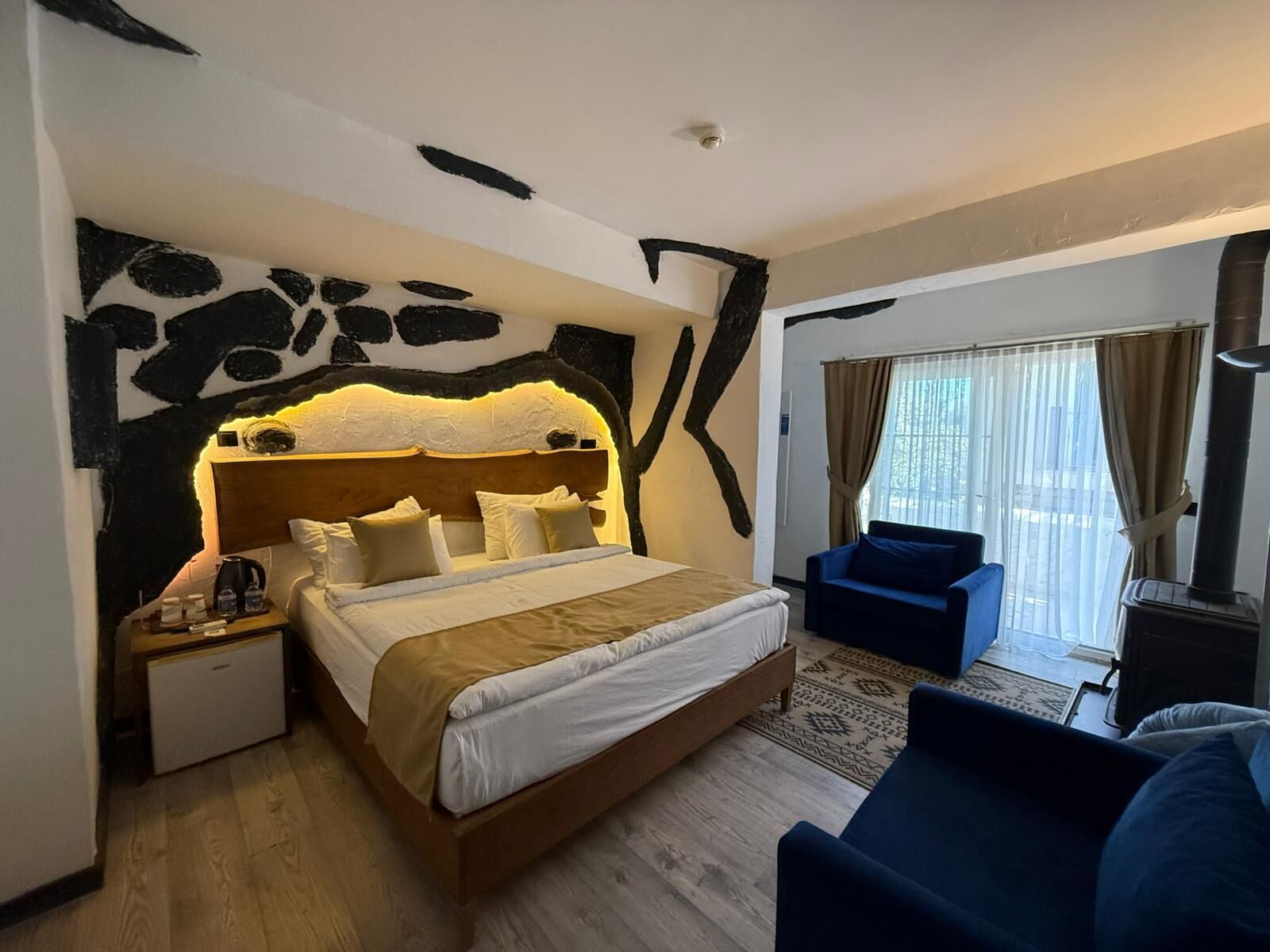 A hotel room with a bed, a nightstand, a blue armchair, a sofa, a window with curtains, and a fireplace.