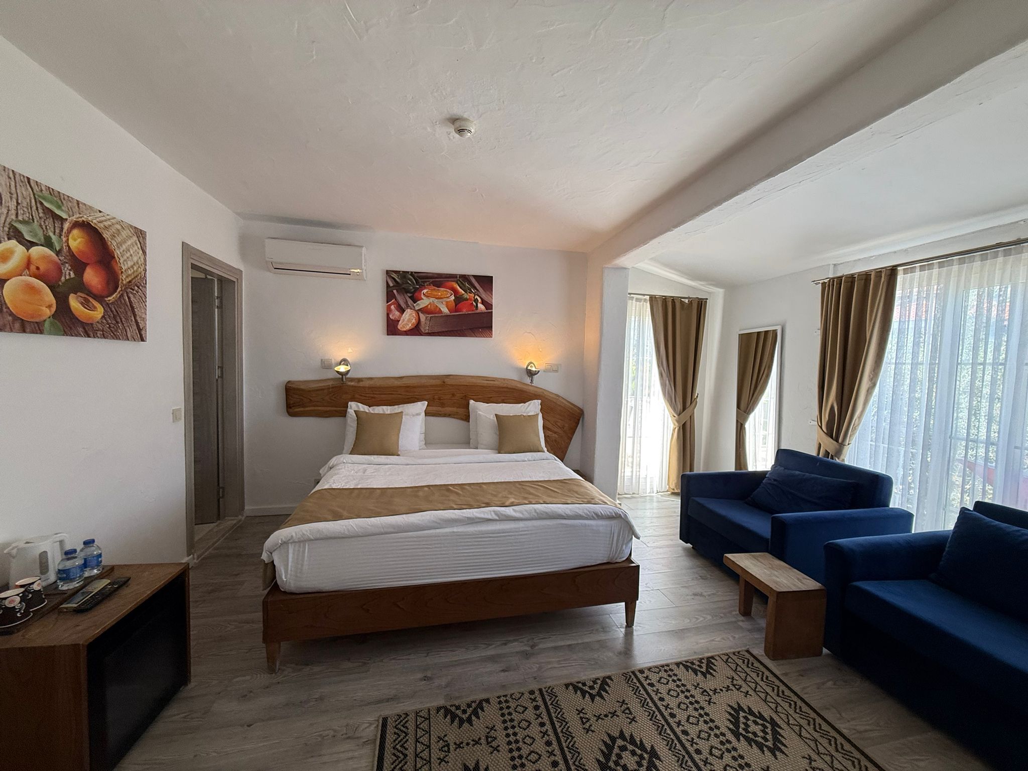 A hotel room with a bed, a blue sofa, a wooden bedside table, a patterned rug, and a fruit-themed painting on the wall.