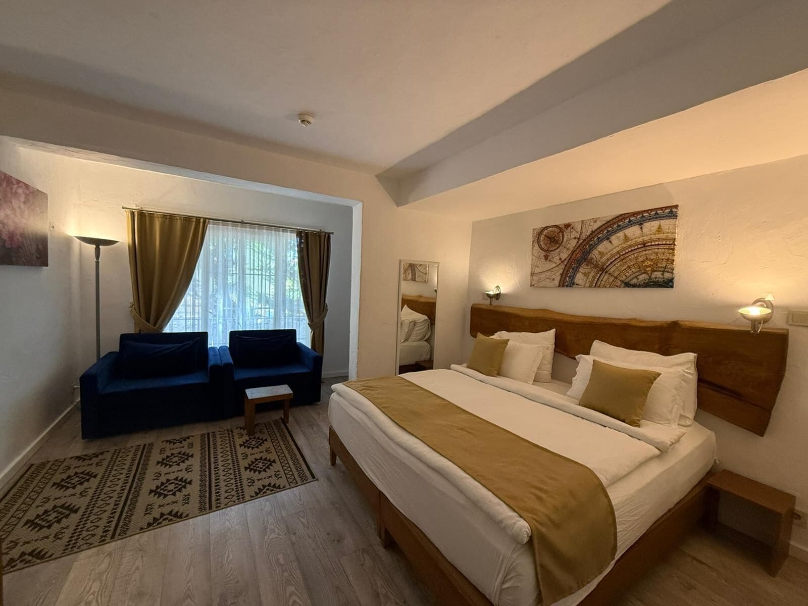 A hotel room with a bed, a sofa, a window with curtains, a floor lamp, and a painting on the wall.