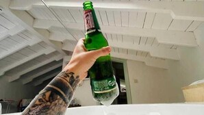 A person holding a bottle of Carlsberg beer in a room with a sloped ceiling and a skylight. - Alaçatı Otel Rebetiko (Cesme)