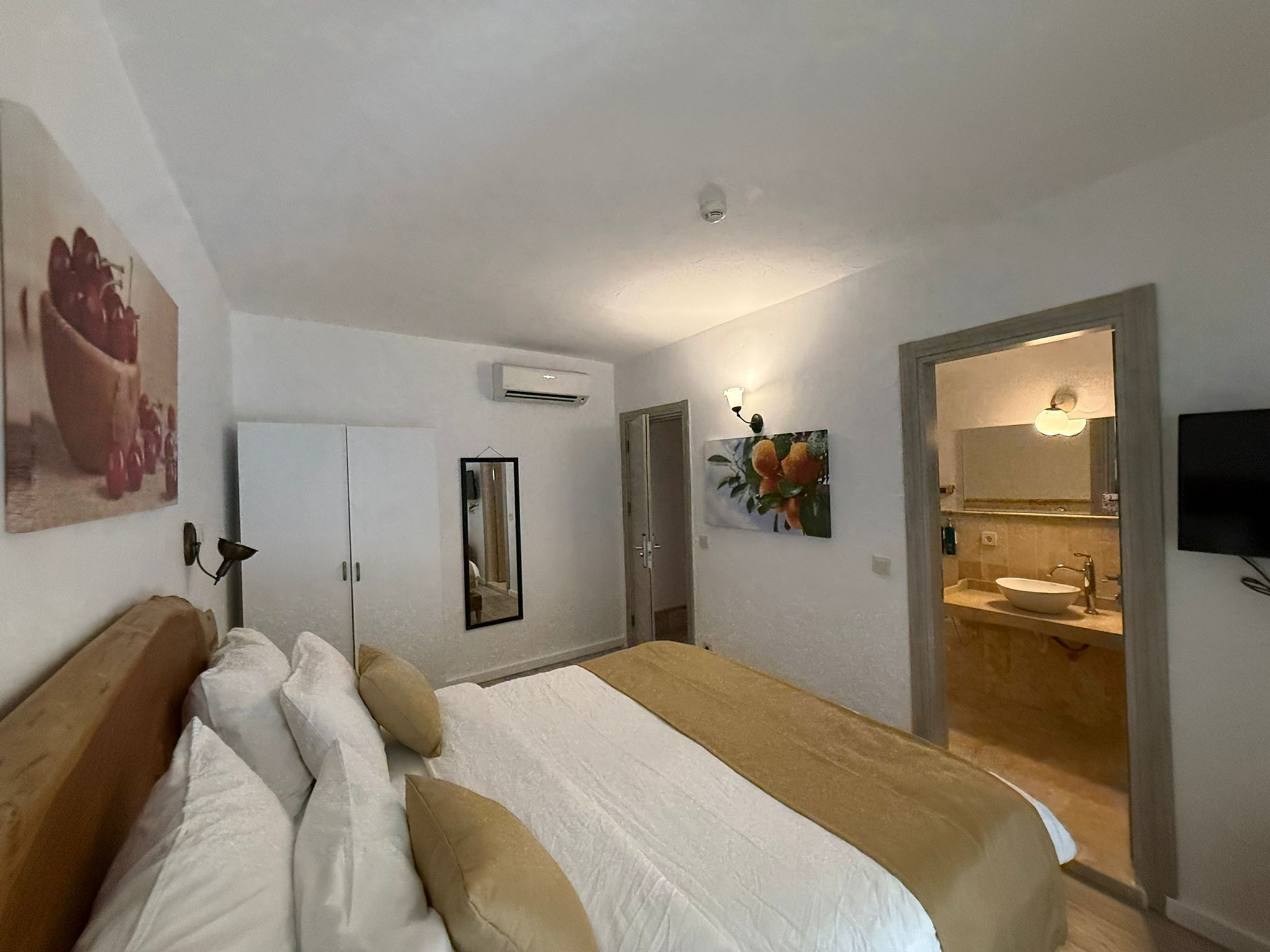 Un dormitorio con cama, armario, espejo, equipo de aire acondicionado y baño con lavabo y espejo.